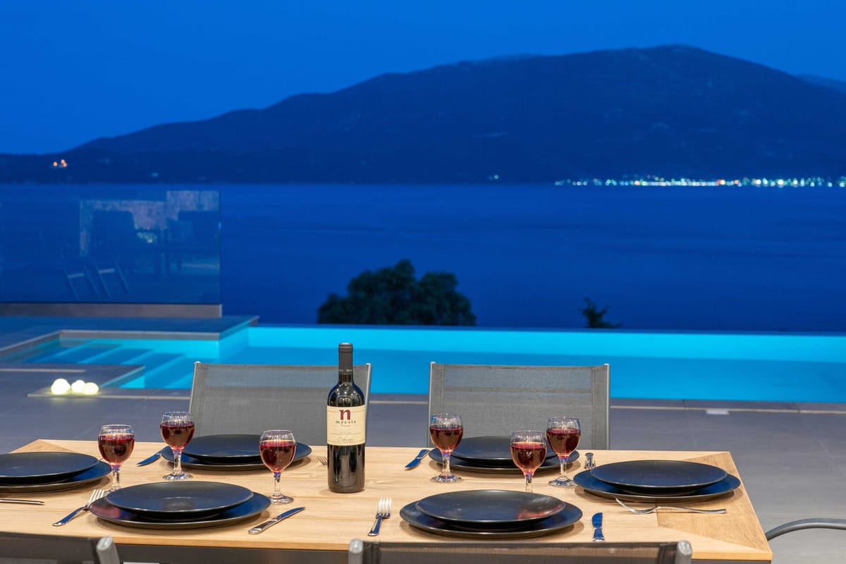 An outdoor dining table set for six at night. Black plates, steel cutlery, and red-wine glasses are arranged on a pale wood table, with a bottle of Myesis red wine at the centre. Behind the table, a glass-sided infinity pool glows blue, and beyond it the dark sea reflects distant town lights. Mountains are silhouetted against a deep-blue dusk sky.