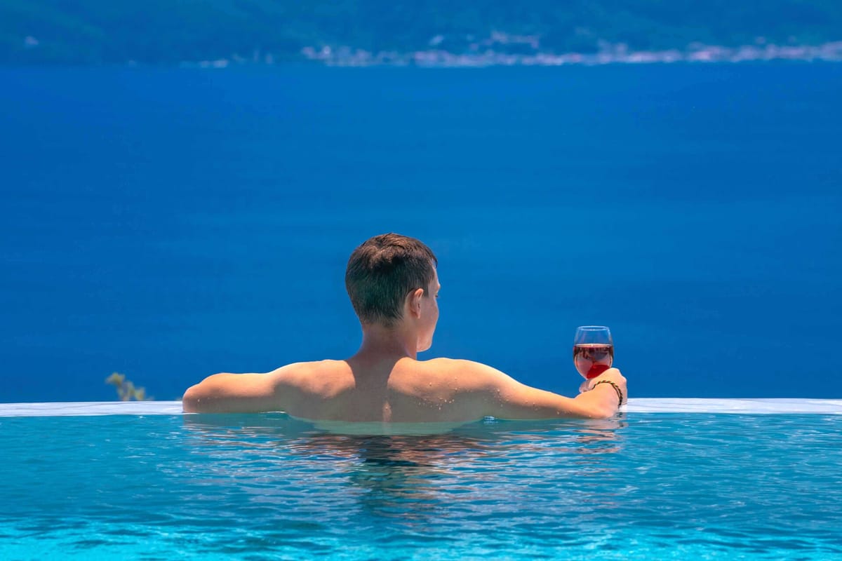 A person seen from behind, shoulders at water level at the edge of an infinity pool, holding a glass of red wine in their right hand. The pool water is vivid turquoise. Beyond the infinity edge the deep blue sea fills most of the frame, with a faint distant shoreline visible on the horizon. Bright daylight.