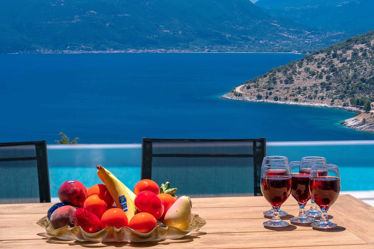 A wooden outdoor table in the foreground holds a shell-shaped bowl filled with fresh fruit — bananas, tomatoes, peaches, and strawberries — alongside four glasses of red wine. Beyond the table, an infinity pool edge is visible, and behind the glass balustrade stretches a sweeping panorama of deep-blue sea, a rocky headland with a coastal road, and mountains on the far shore under bright midday sun.