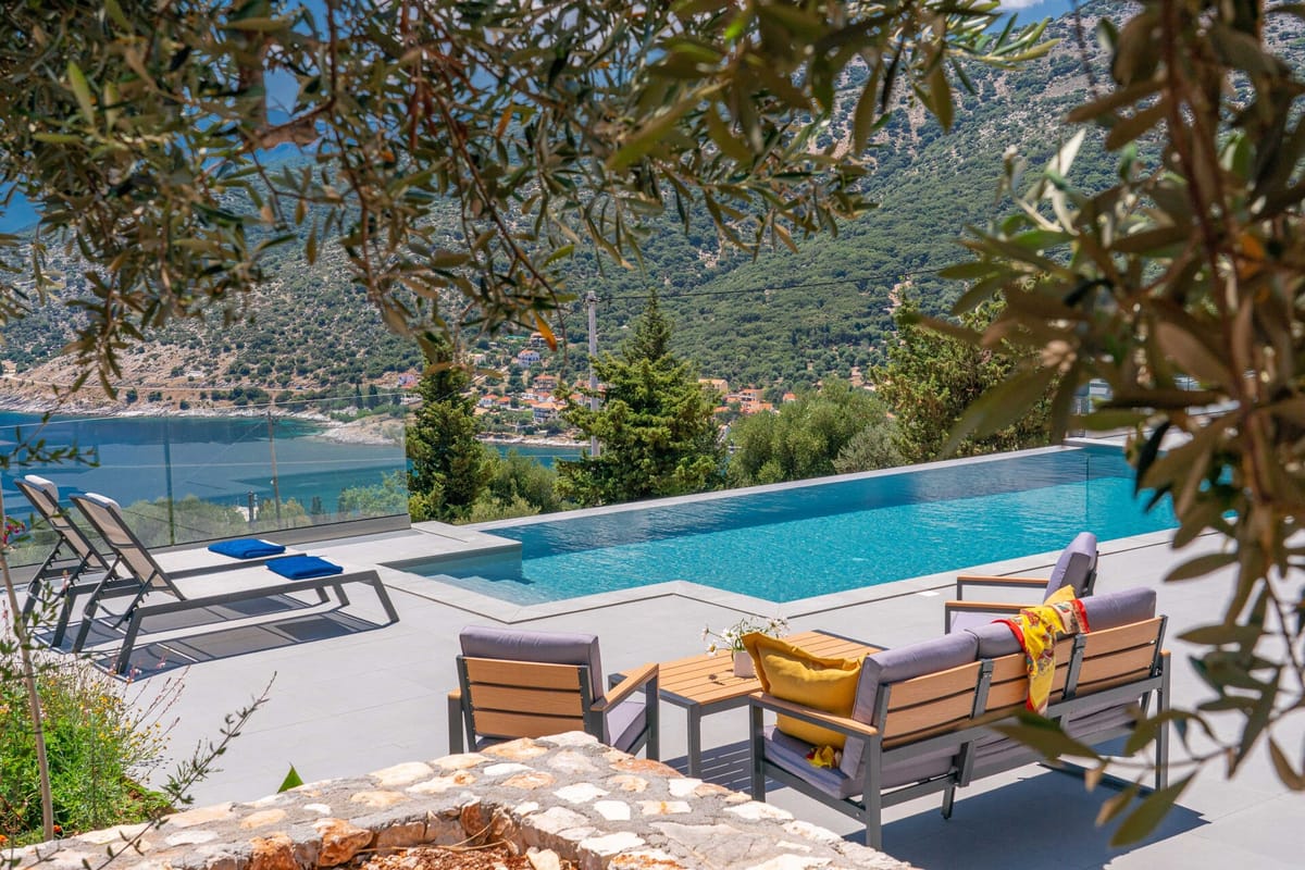A rectangular infinity pool with a glass balustrade sits on a wide pale-stone terrace. Grey sun loungers with blue towels are arranged to the left. A grey outdoor sofa set with yellow cushions occupies the foreground right. The pool overlooks a hillside village, cypress and olive trees, and a deep-blue bay. An olive tree with silver-green leaves frames the top of the image. Bright daytime light.