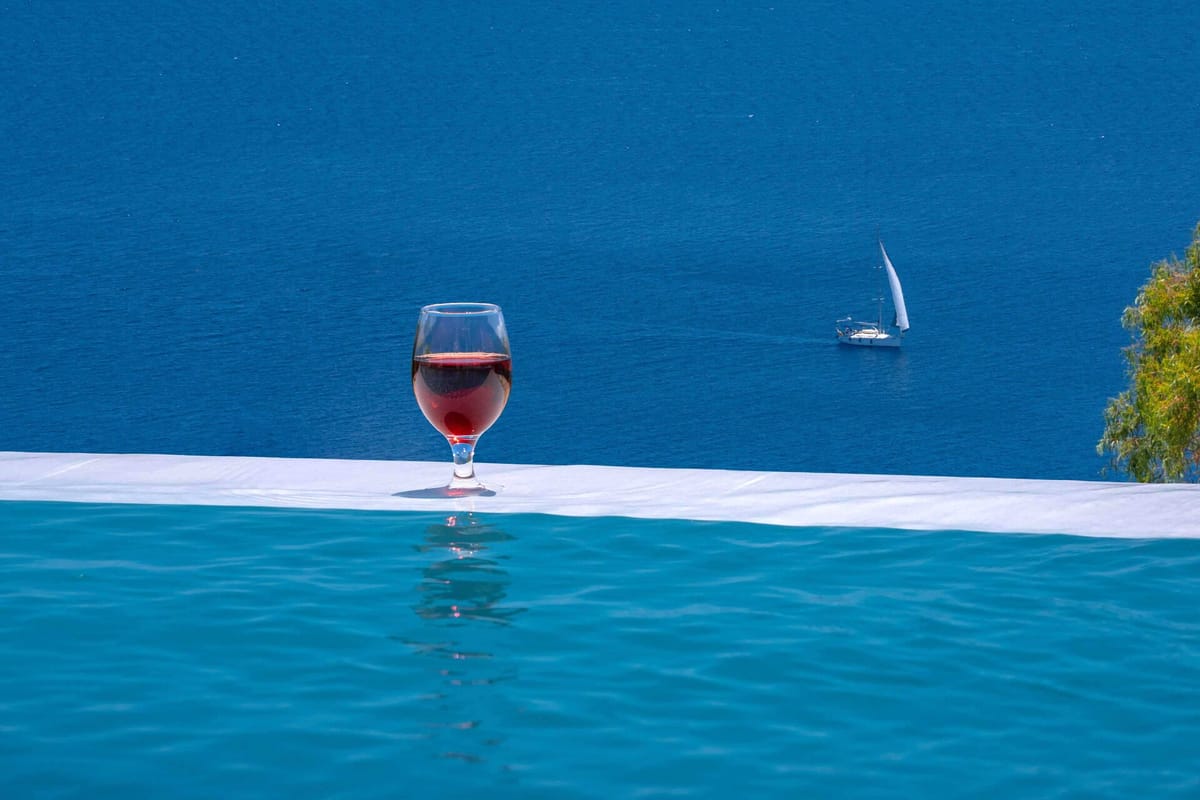 Close-up of a wine glass containing red or rosé wine resting on the white coping of an infinity pool. The turquoise pool water fills the lower half of the frame. Beyond the pool's vanishing edge, a vast deep blue sea dominates the background with a single white sailing yacht visible mid-frame to the right.