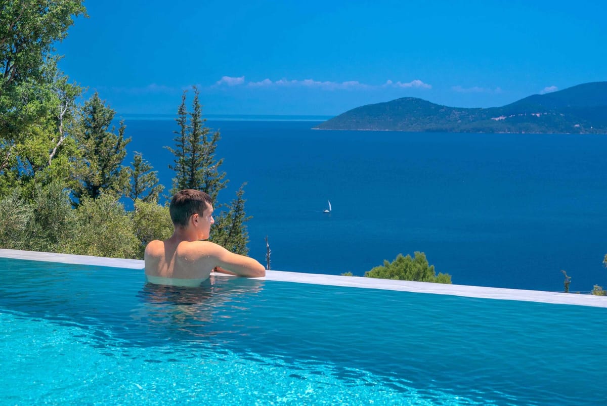 A person from behind resting at the infinity pool edge, looking out over deep-blue open sea with a sailing boat, a wooded island, and tall pine trees, under a bright blue sky.