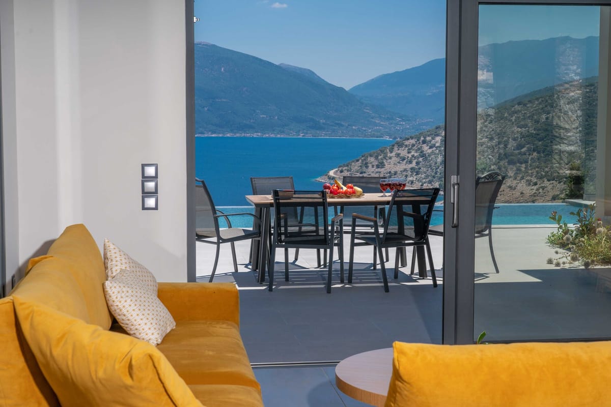 View from inside the living room looking through open sliding glass doors onto a sun-drenched terrace. In the foreground, a mustard-yellow sofa with a patterned cushion is visible on both sides. On the terrace, a rectangular dining table with black chairs holds a fruit bowl and two glasses of red wine. Beyond the terrace, a pool edge is just visible to the right, and a sweeping panorama of vivid blue sea flanked by two rugged mountain ranges stretches under a bright midday sky.