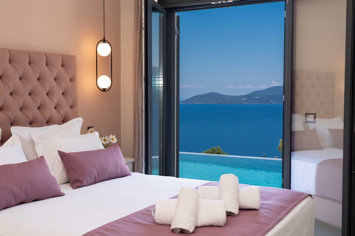 Double bedroom with a large tufted blush-pink upholstered headboard, white bedding, and two mauve accent pillows. Two rolled white towels are placed on the bed. A double-globe pendant light in a black oval frame hangs to the left of the bed. Floor-to-ceiling bi-fold glass doors are open to reveal a turquoise infinity pool in the foreground and a deep-blue sea with a green-forested island or headland under a vivid blue sky beyond. The walls are a warm sandy beige. Small white daisy flowers are visible on the bedside table.
