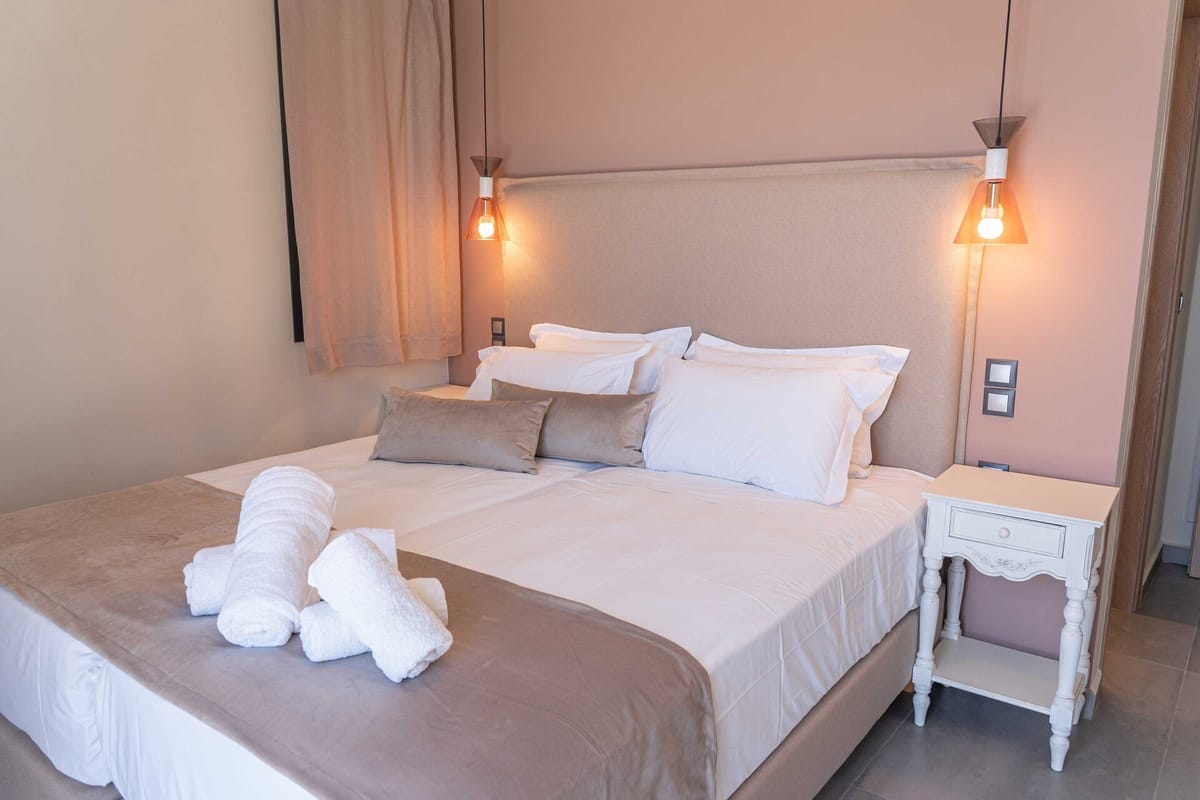 A double bedroom with a large upholstered fabric headboard in beige-grey, dressed with white hotel-style pillows and grey velvet rectangular cushions, a taupe velvet bedspread with two rolled white towels placed at the foot of the bed, two pendant lights with amber conical shades hanging either side of the headboard, a white ornate bedside table with a single drawer visible on the right, linen curtains in a warm blush-pink tone, and walls painted in a warm dusty rose.