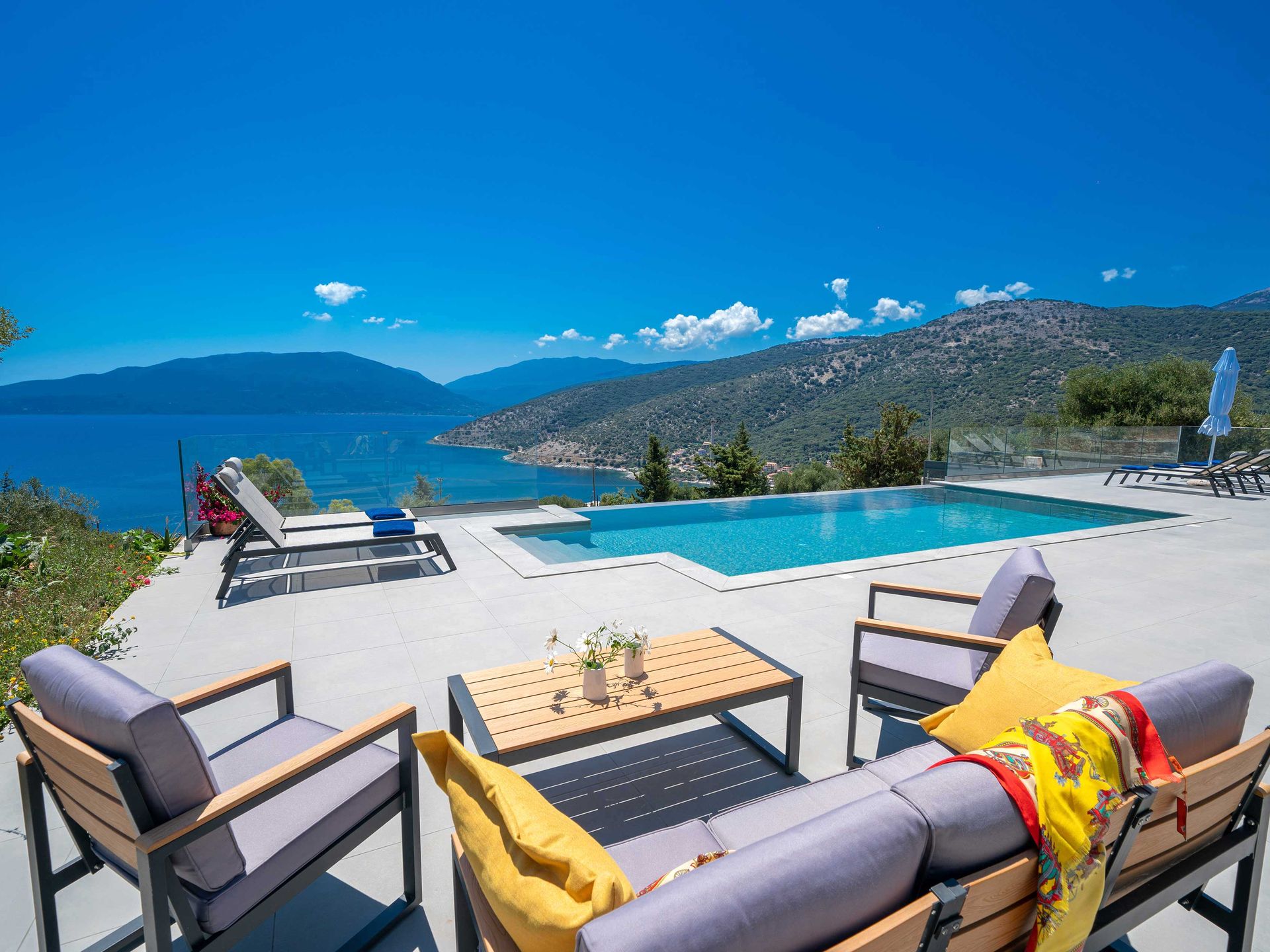 Daytime poolside terrace with infinity-edge pool overlooking a mountainous coastline and bay. Grey outdoor lounge furniture with grey cushions and yellow and red patterned throw pillows sits in the foreground around a wooden coffee table with a small white flower arrangement. Sun loungers and a closed umbrella are visible to the right, and a lounger with red flowers to the left, behind a frameless glass balustrade.